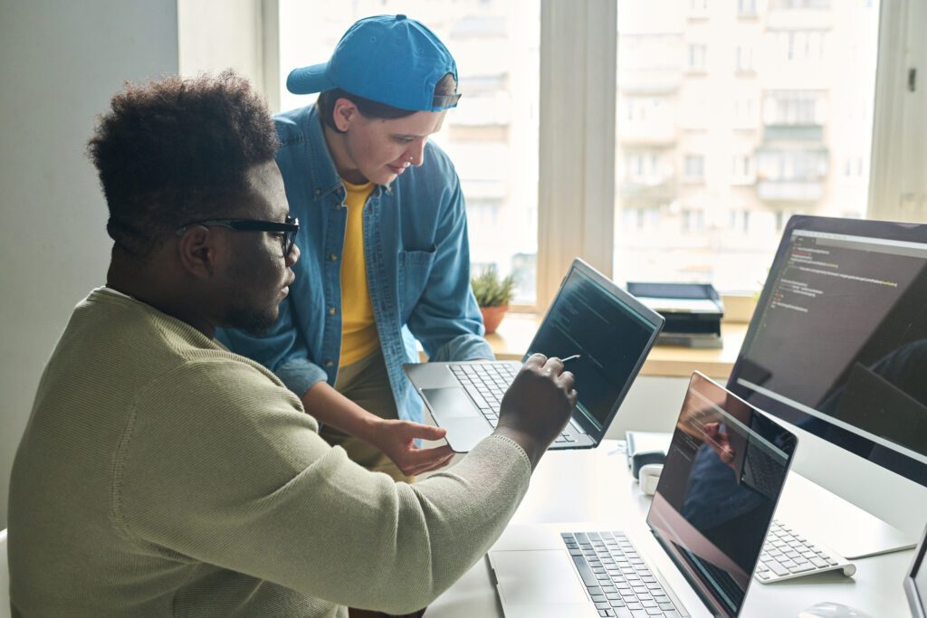 African young programmer in eyeglasses pointing at software on screen of laptop and consulting with his colleague